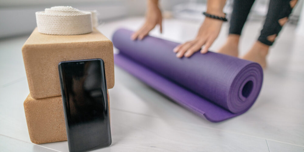 Phone app screen closeup at fitness yoga studio in indoor gym or at home. Girl rolling out mat getting ready for fit exercise workout with mobile smartphone showing online class.